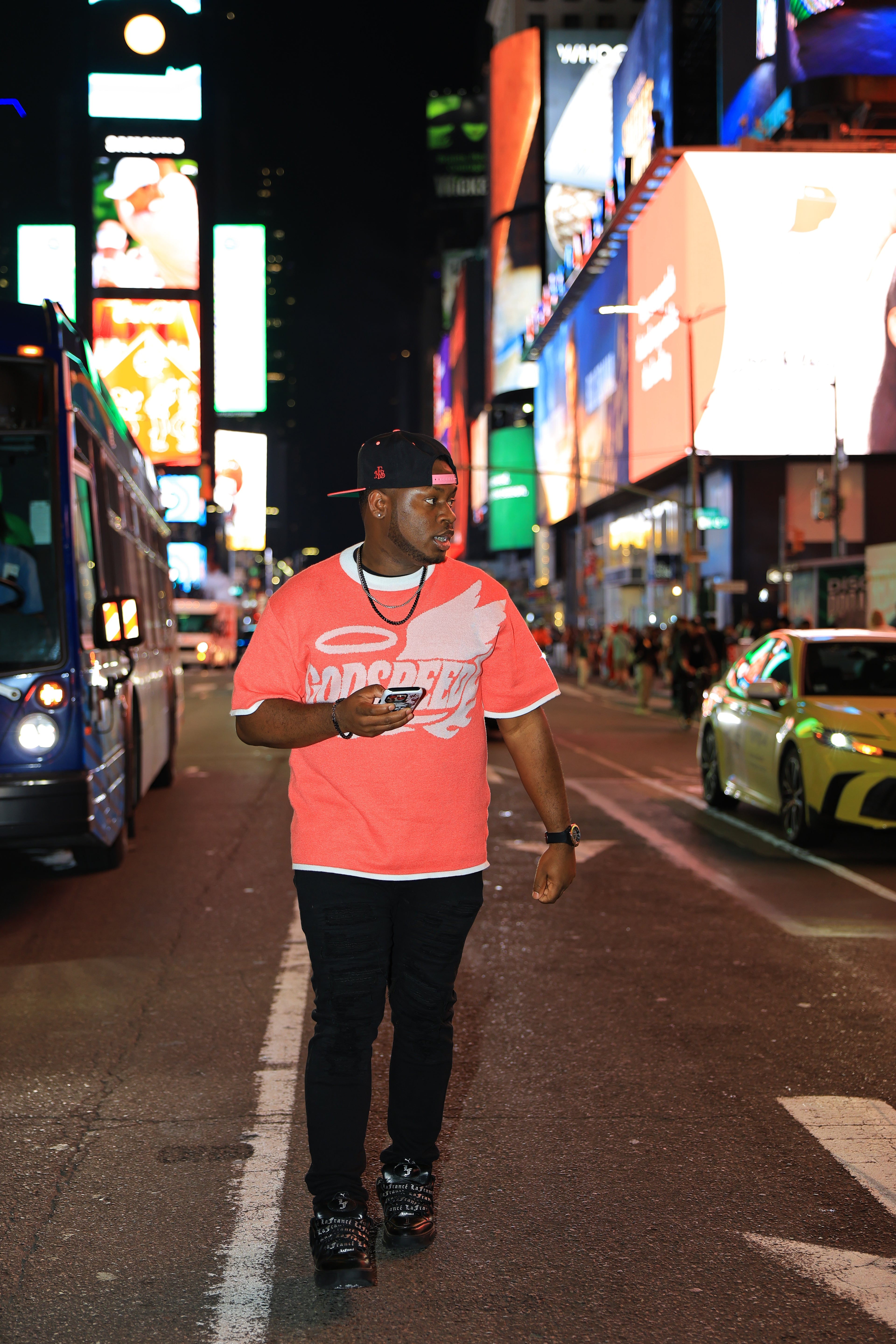 Man in a bright pink shirt walking on a city street at night with illuminated billboards in the background.